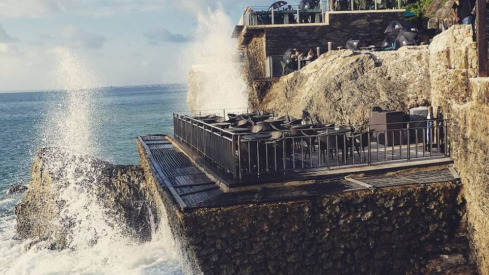 A coastal cliff with a rocky platform overlooking the sea, featuring a restaurant and a stone wall.