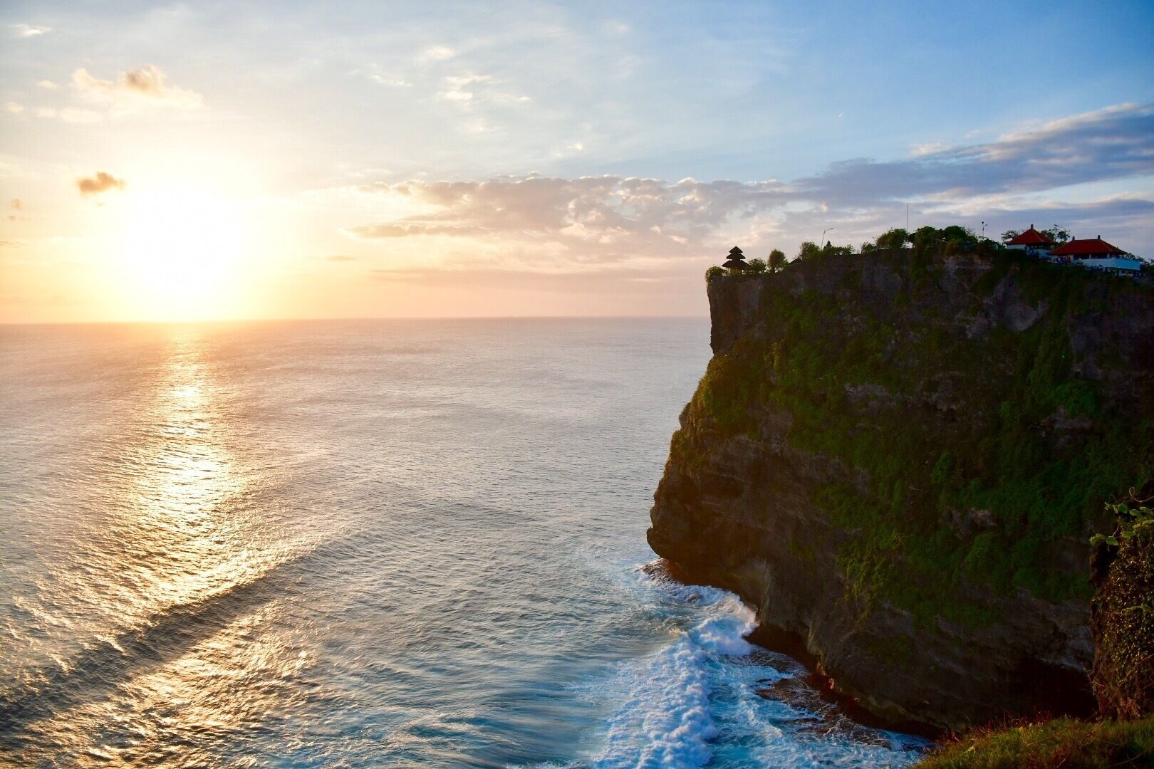 A coastal cliff with a sunset, ocean waves, and a pagoda on top.