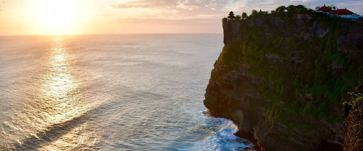A coastal cliff with a sunset, ocean waves, and a pagoda on top.
