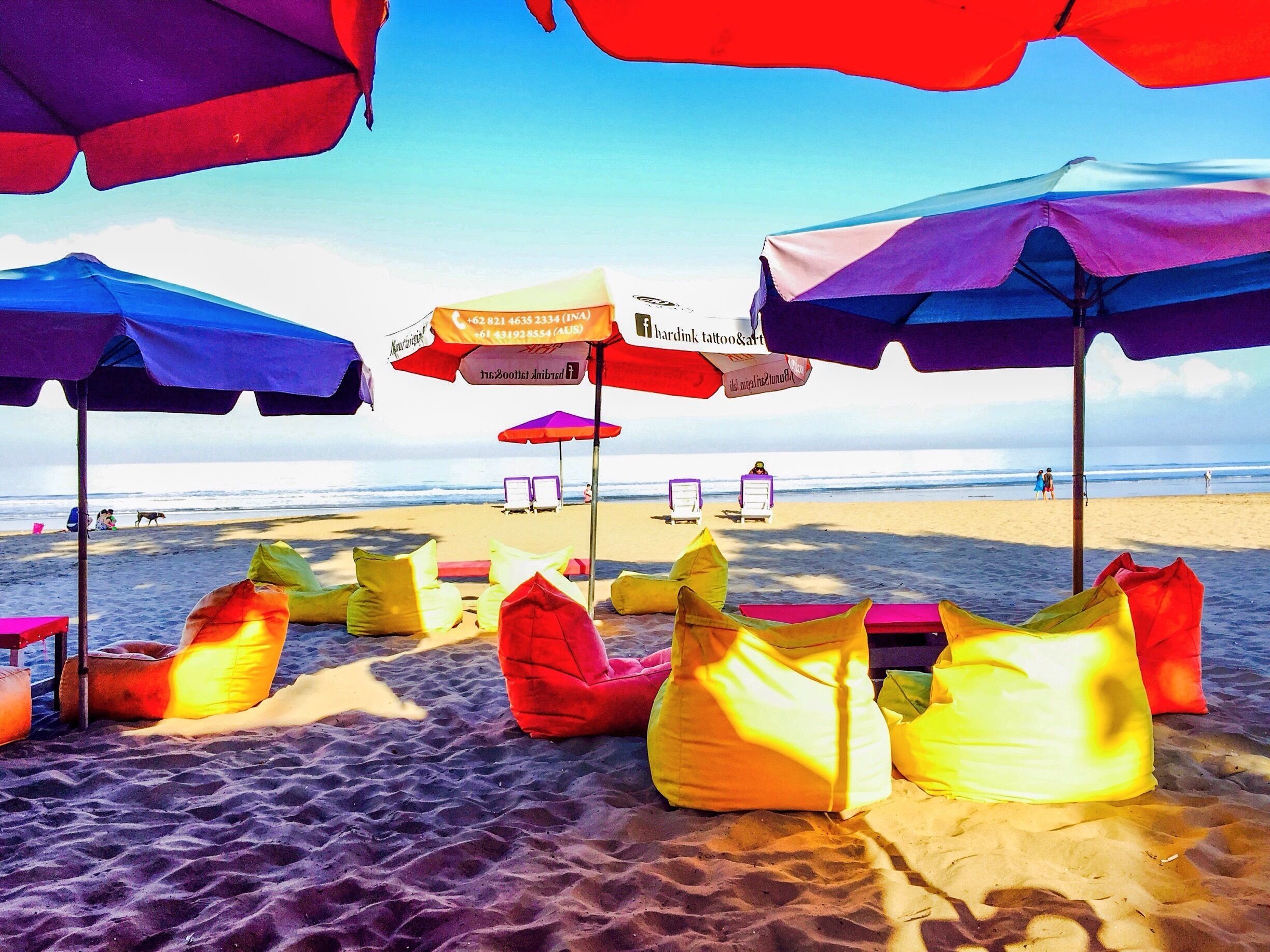 Sombrillas de playa coloridas y pufs sobre arena, con el océano al fondo.