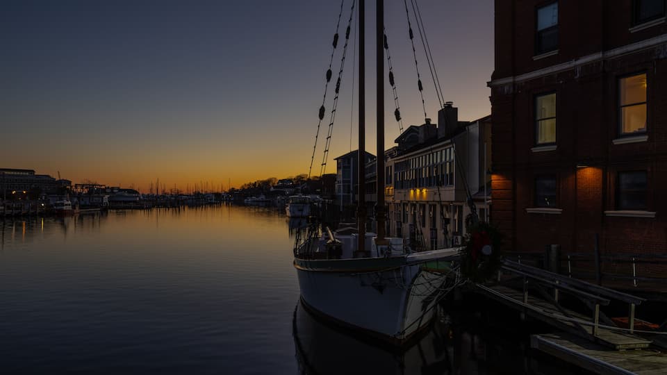 A calm harbor at dusk with a boat docked on the left and buildings along the right.
