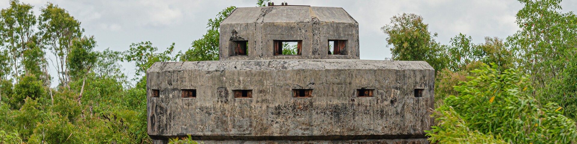 Una construcción de concreto erosionado con varias ventanas y una pequeña torre en la parte superior, rodeada de abundante vegetación.