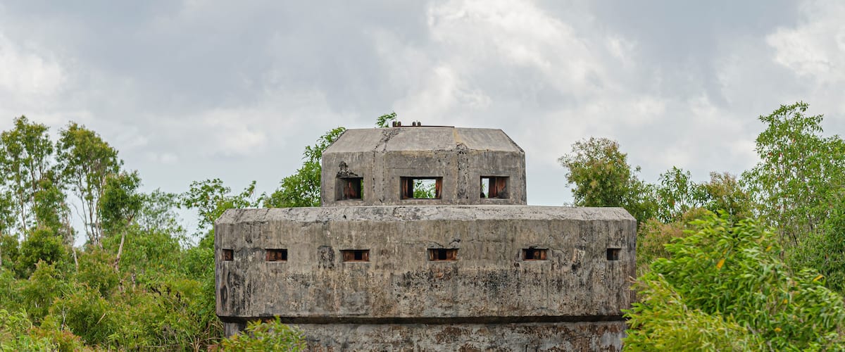 A weathered concrete structure with multiple windows and a small tower on top, surrounded by dense greenery.