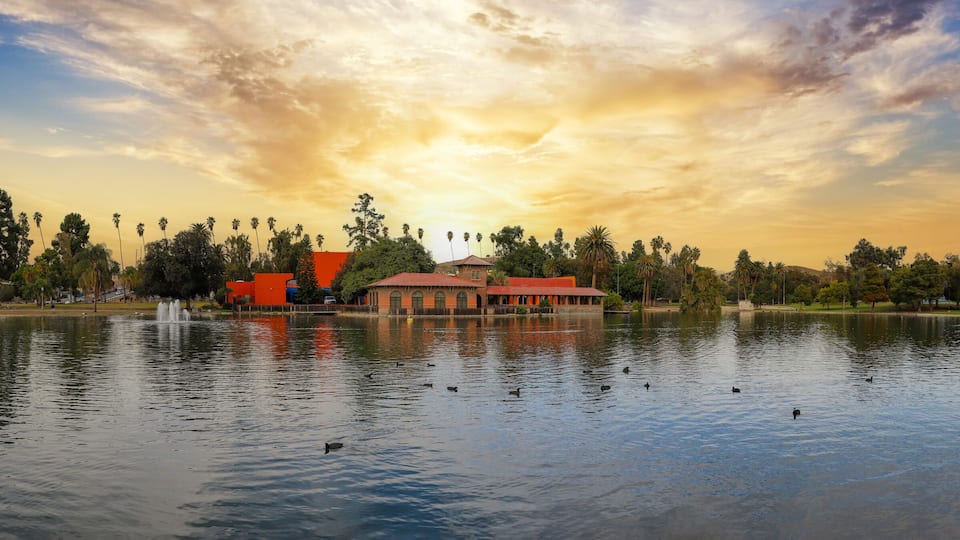 a gorgeous autumn landscape at Lincoln Park with a lake surrounded by lush green palm trees and plants, birds swimming in the water, buildings, powerful clouds at sunset in Los Angeles California USA