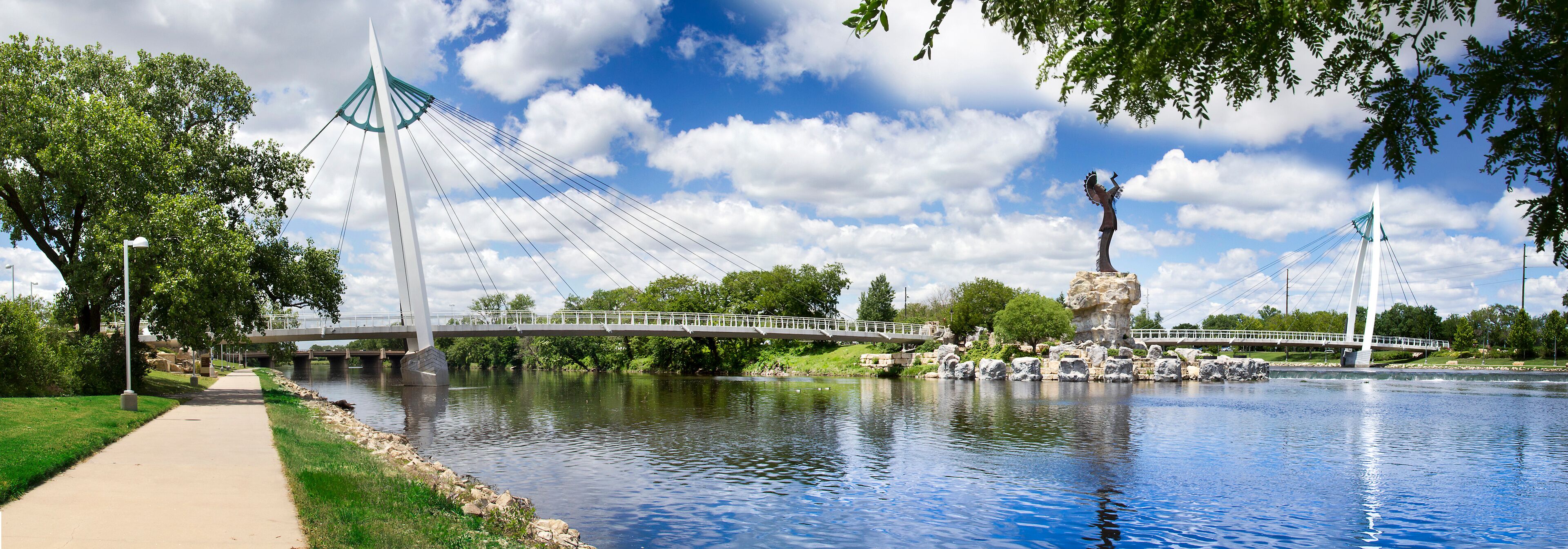 A river with a bridge, a statue, and a park with trees and a walkway.