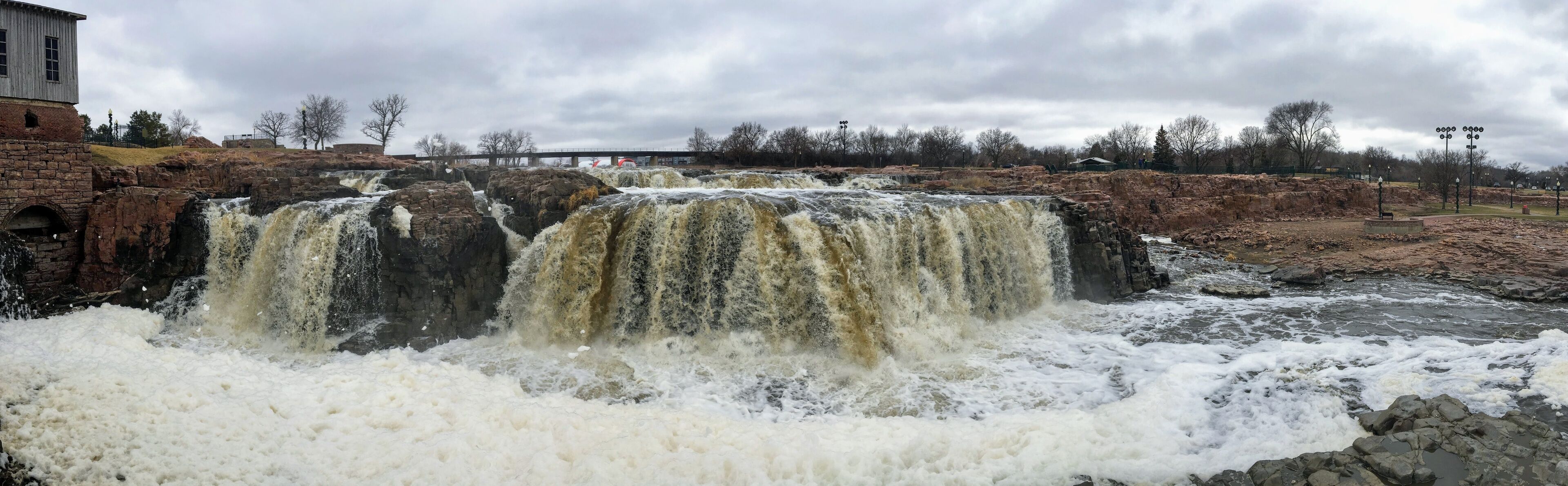 The Big Sioux River flows over rocks in Sioux Falls South Dakota with views of wildlife, ruins, park paths, train track bridge, trees and city in the surrounding area and background