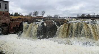 The Big Sioux River flows over rocks in Sioux Falls South Dakota with views of wildlife, ruins, park paths, train track bridge, trees and city in the surrounding area and background