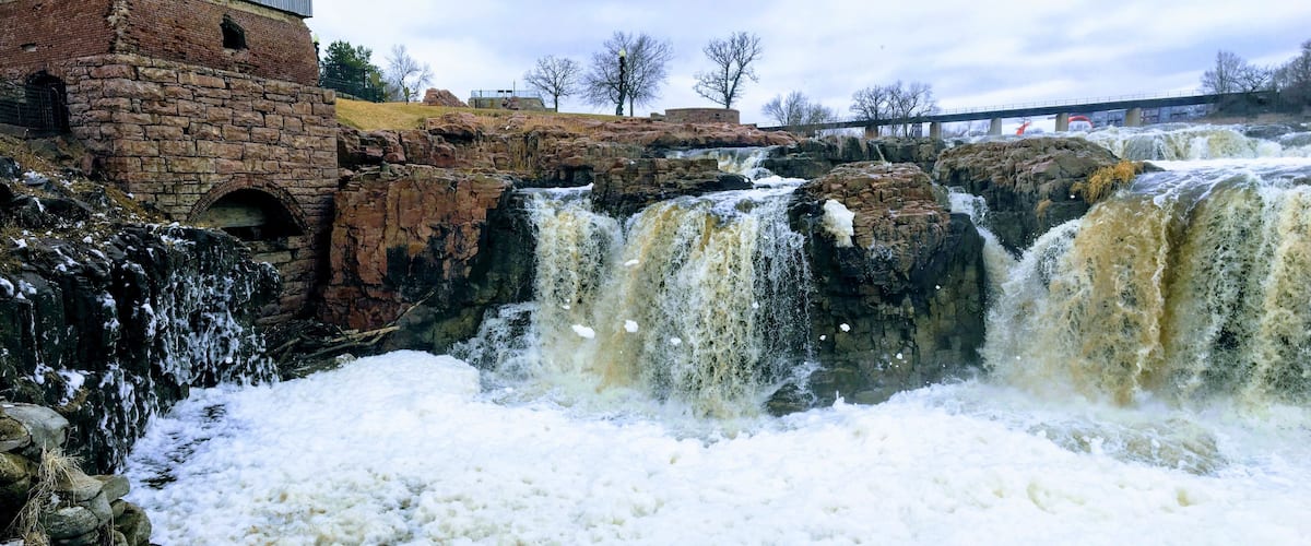 The Big Sioux River flows over rocks in Sioux Falls South Dakota with views of wildlife, ruins, park paths, train track bridge, trees and city in the surrounding area and background