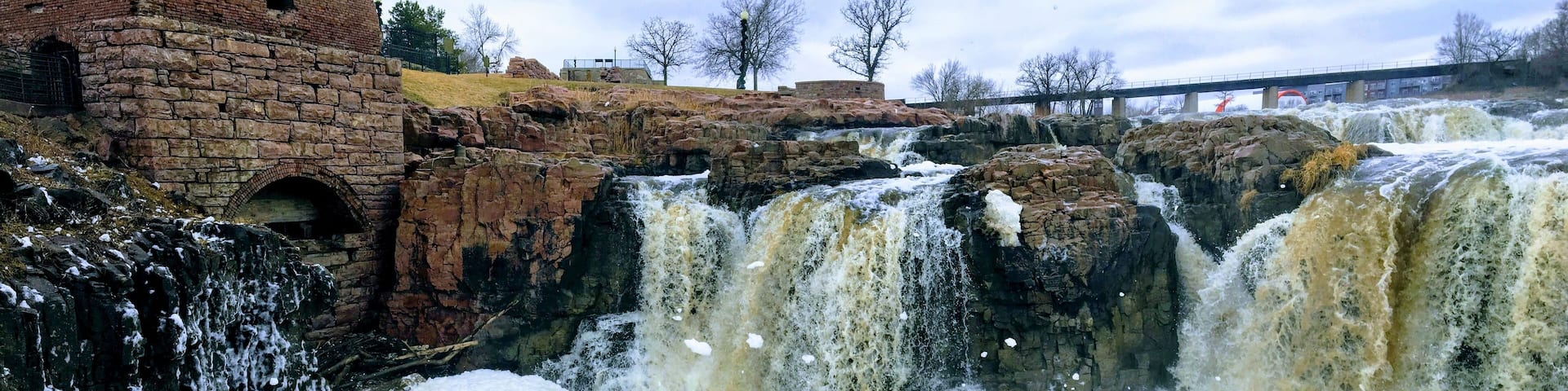 The Big Sioux River flows over rocks in Sioux Falls South Dakota with views of wildlife, ruins, park paths, train track bridge, trees and city in the surrounding area and background