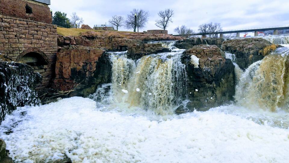 The Big Sioux River flows over rocks in Sioux Falls South Dakota with views of wildlife, ruins, park paths, train track bridge, trees and city in the surrounding area and background