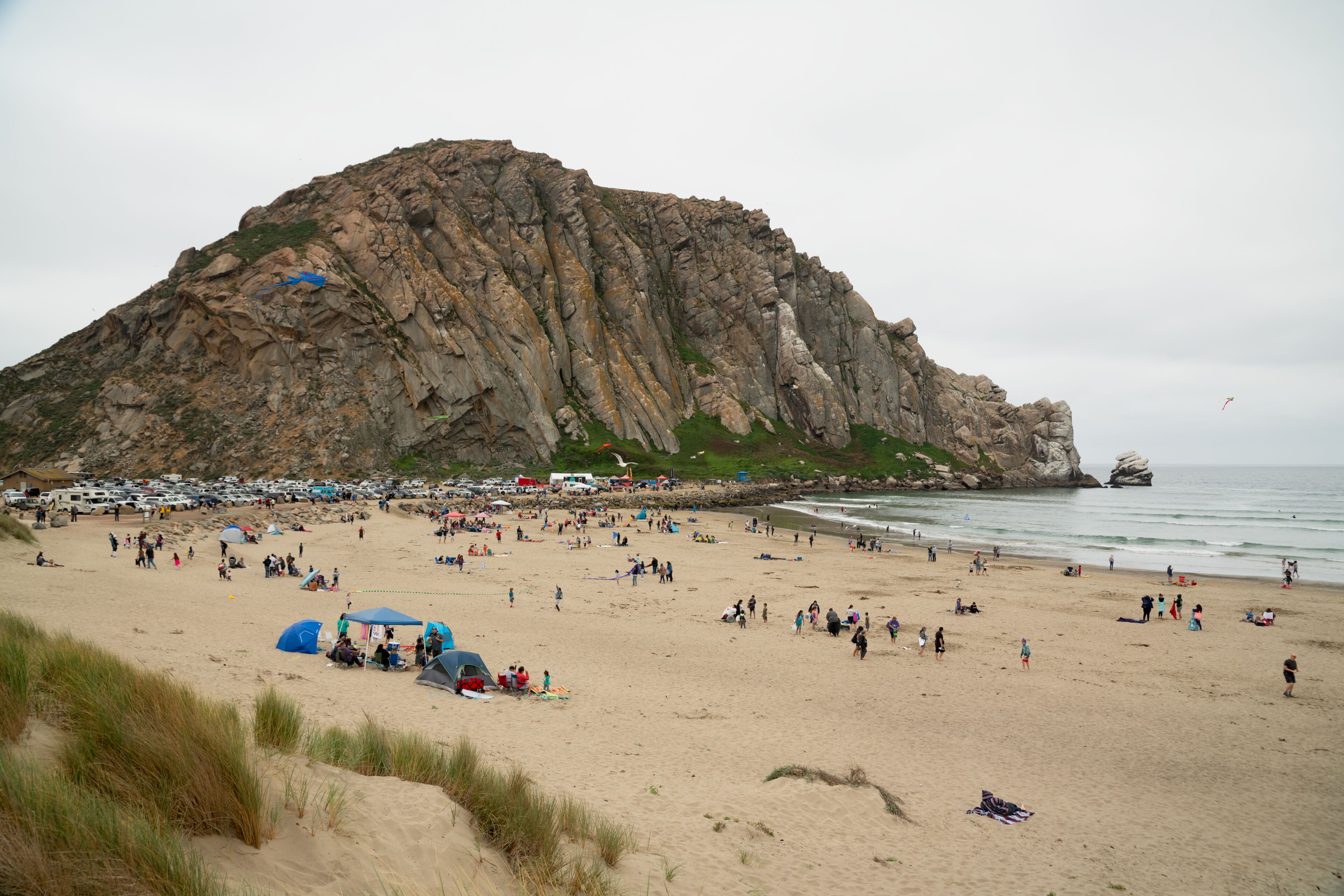 Uma praia com uma grande colina rochosa, tendas e pessoas.