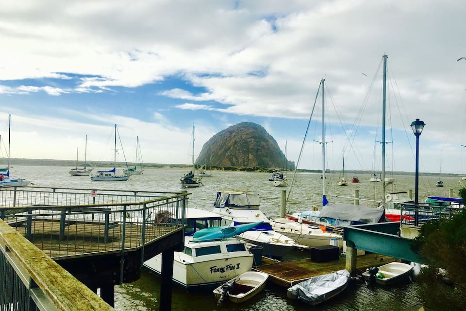 Een jachthaven met diverse boten afgemeerd, een houten steiger en een opvallend rotsachtig eiland op de achtergrond.