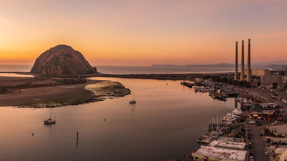 A coastal town with a prominent rock formation, a marina with boats, and industrial chimneys in the background.