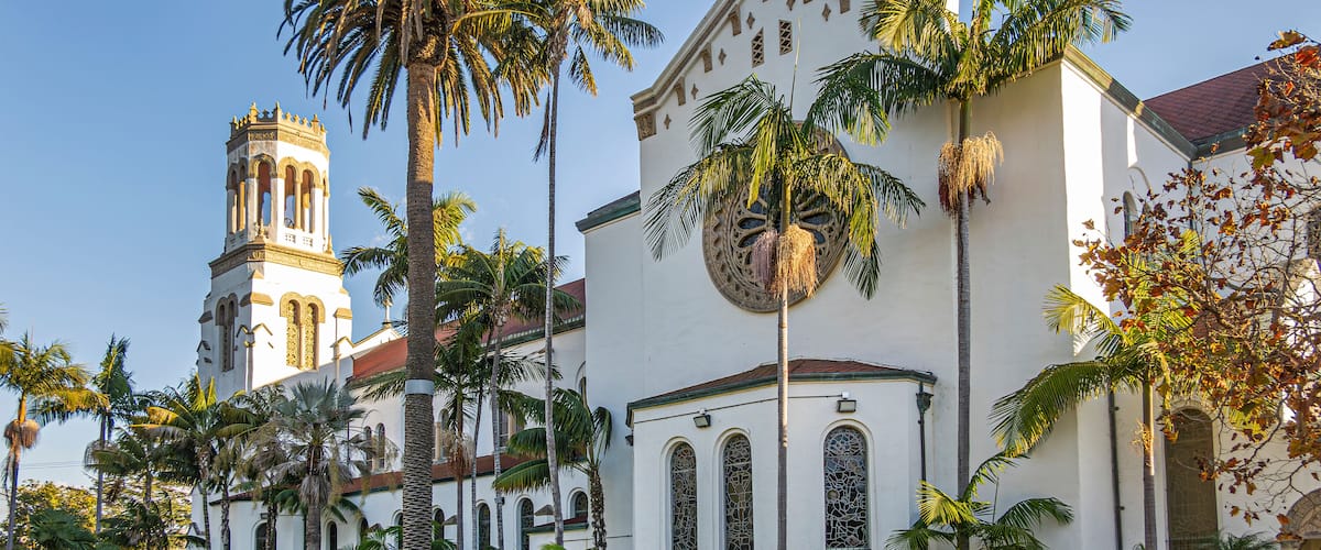 Santa Barbara, CA, USA - January 19, 2024: Beige west facade of Our Lady of Sorrows church with sunlight  on tower against blue sky. Green palm tree  foliage and lawn in front.