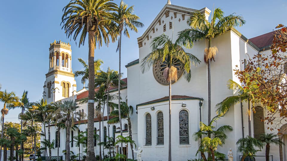 Santa Barbara, CA, USA - January 19, 2024: Beige west facade of Our Lady of Sorrows church with sunlight on tower against blue sky. Green palm tree foliage and lawn in front.