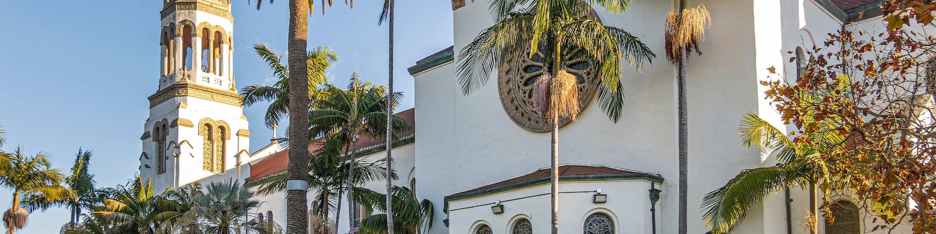 Santa Barbara, CA, USA - January 19, 2024: Beige west facade of Our Lady of Sorrows church with sunlight on tower against blue sky. Green palm tree foliage and lawn in front.