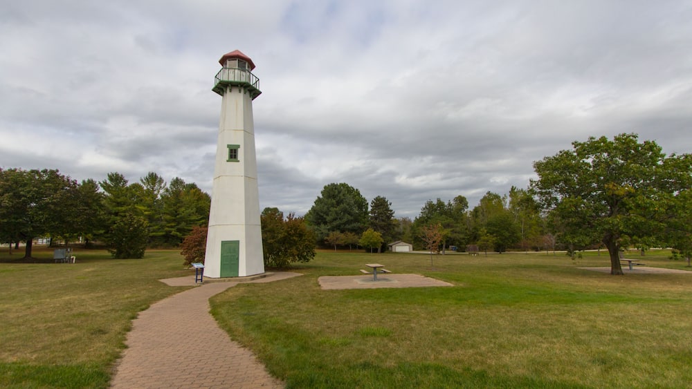 Un phare blanc, doté d’une porte verte et d’un toit rouge, est entouré d’un parc verdoyant avec des arbres et un sentier dallé.