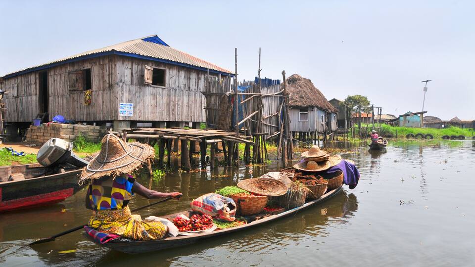 A wooden house with a blue roof, a thatched-roof house, and a wooden dock extending into the water.
