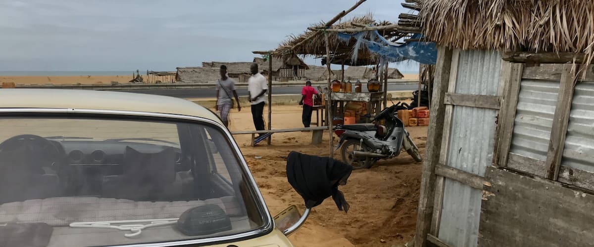A beige car parked near a sandy area with a thatched-roof structure and a motorcycle.