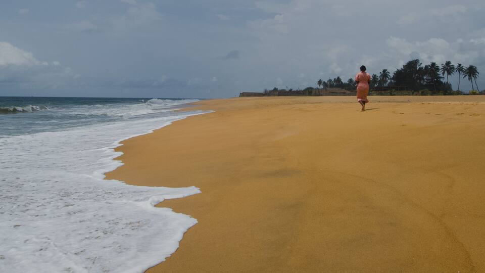 A sandy beach with waves, a person walking, and palm trees in the background.