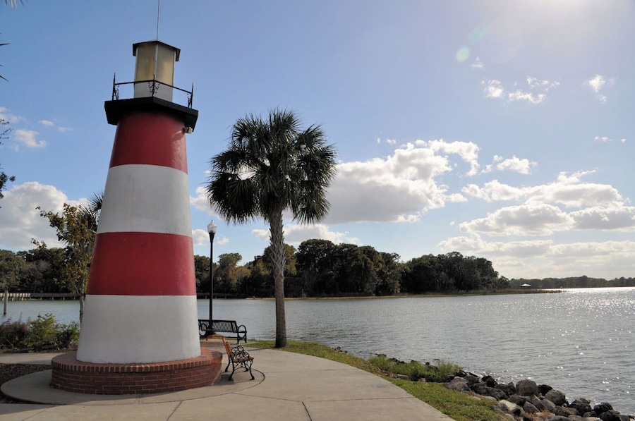 Un phare rouge et blanc se dresse au bord d’un lac paisible, agrémenté de bancs et de palmiers.