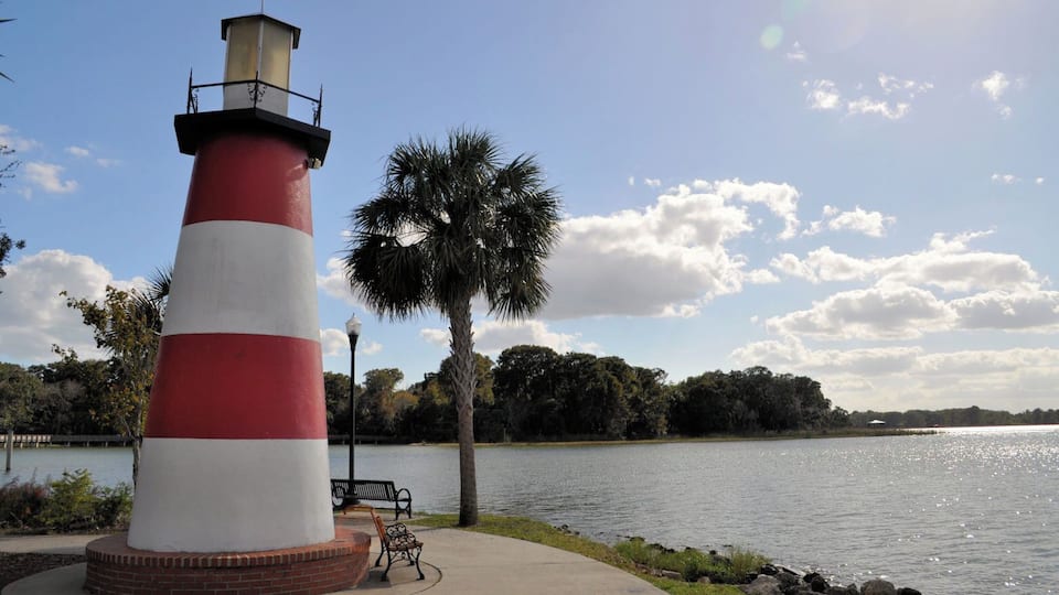 A red and white lighthouse by a calm lake with benches and palm trees.
