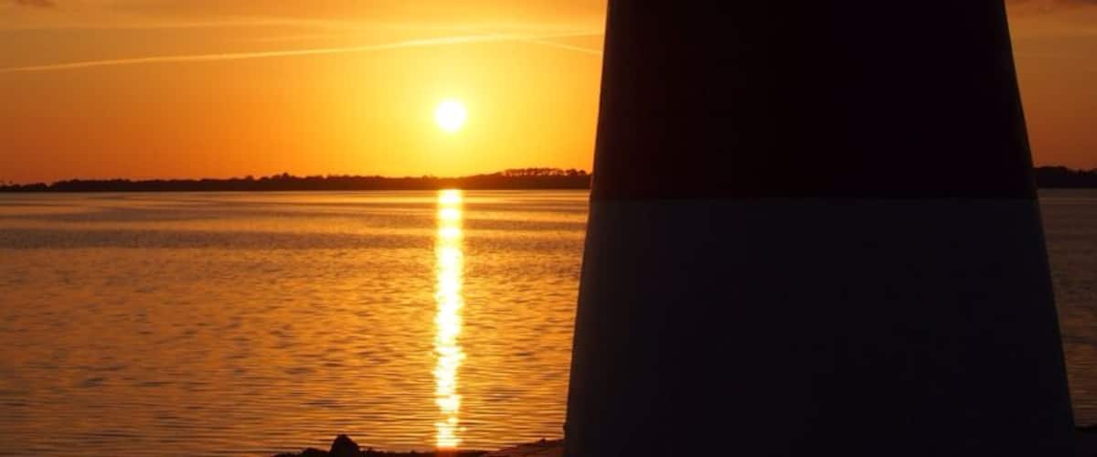 A lighthouse at sunset with the sun reflecting on the water.
