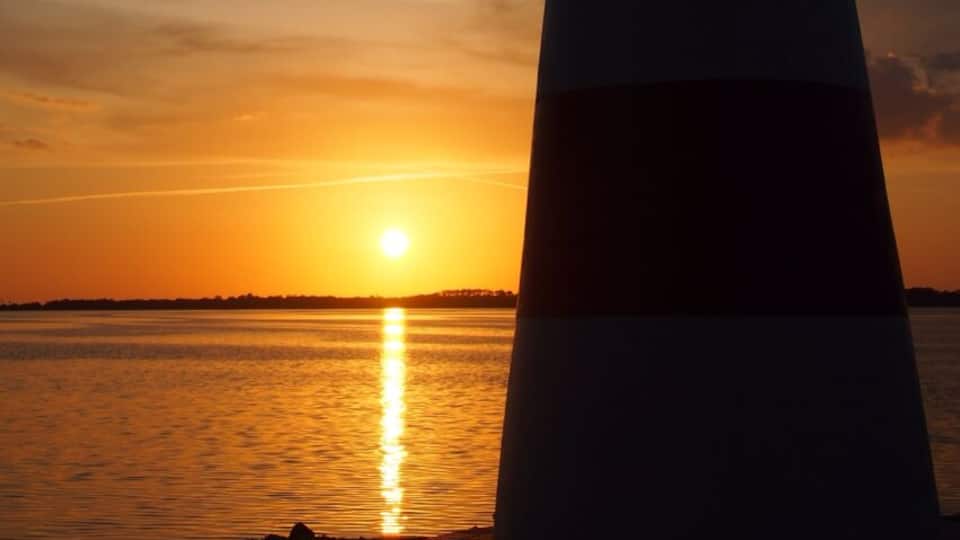 A lighthouse at sunset with the sun reflecting on the water.