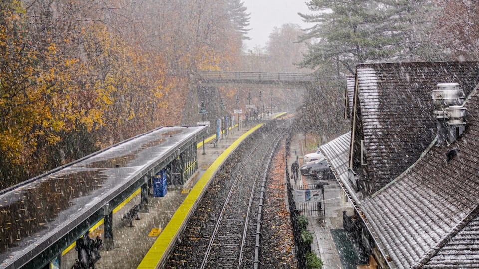 A train station with tracks, a platform, and trees with autumn leaves.