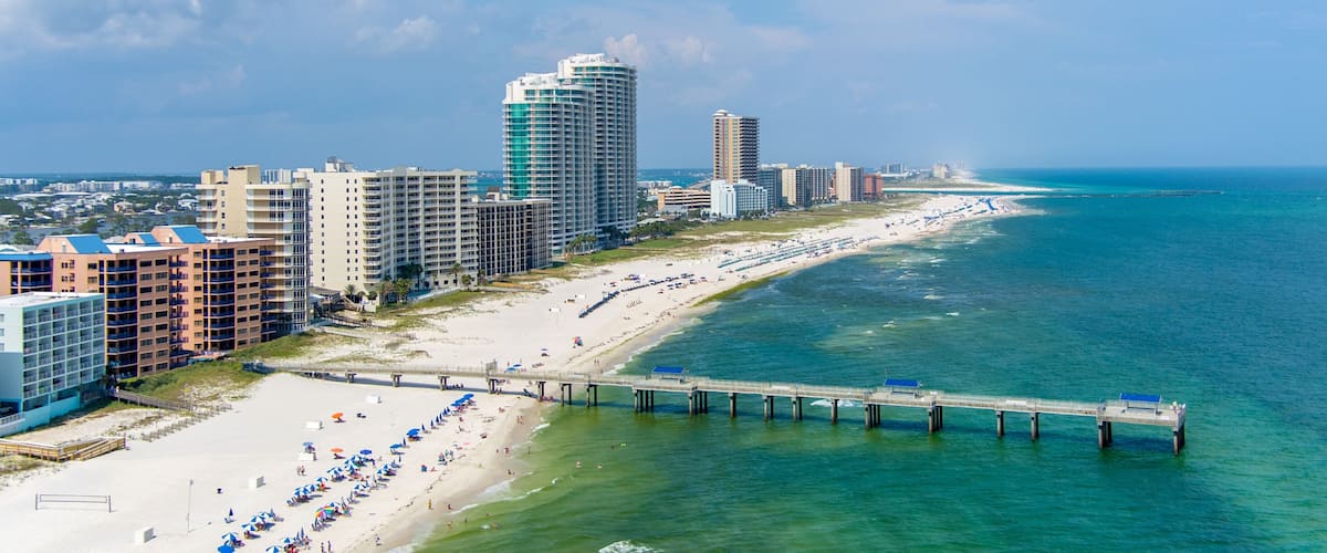 A coastal cityscape with a long sandy beach, a pier extending into the ocean, and high-rise buildings.
