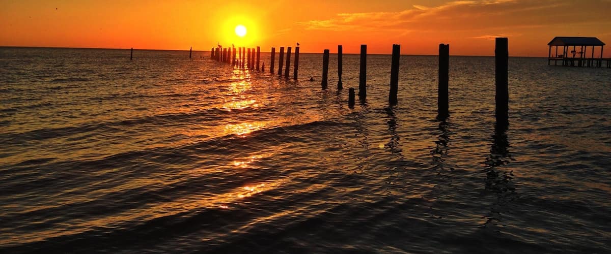 A sunset over a calm sea with wooden posts and a small structure on the right.