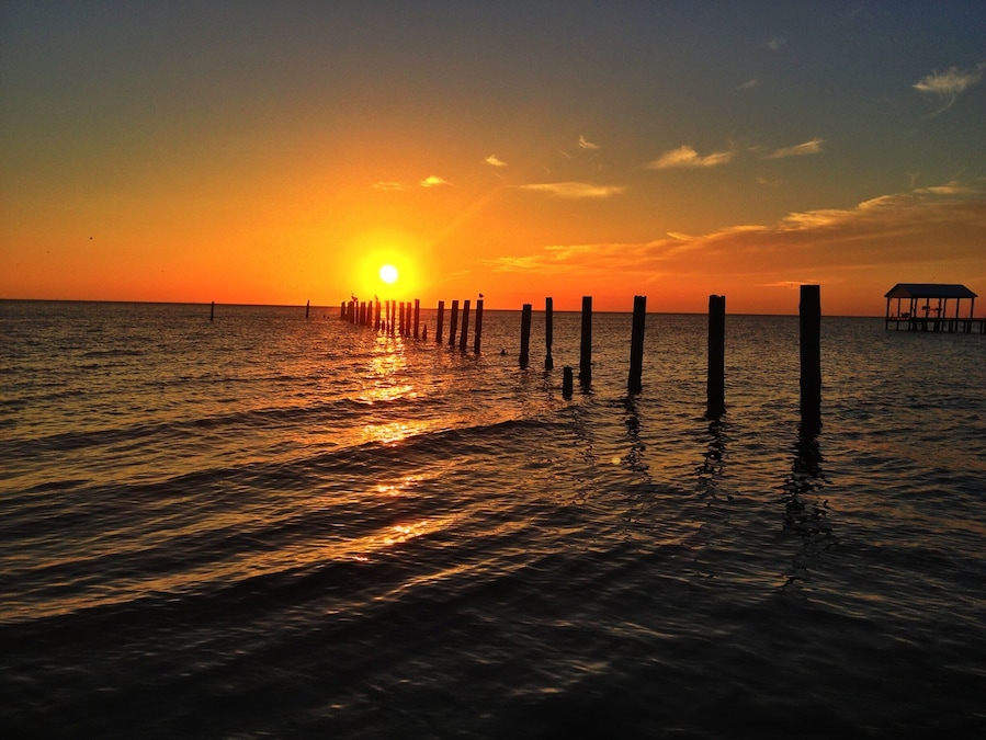 A sunset over a calm sea with wooden posts and a small structure on the right.