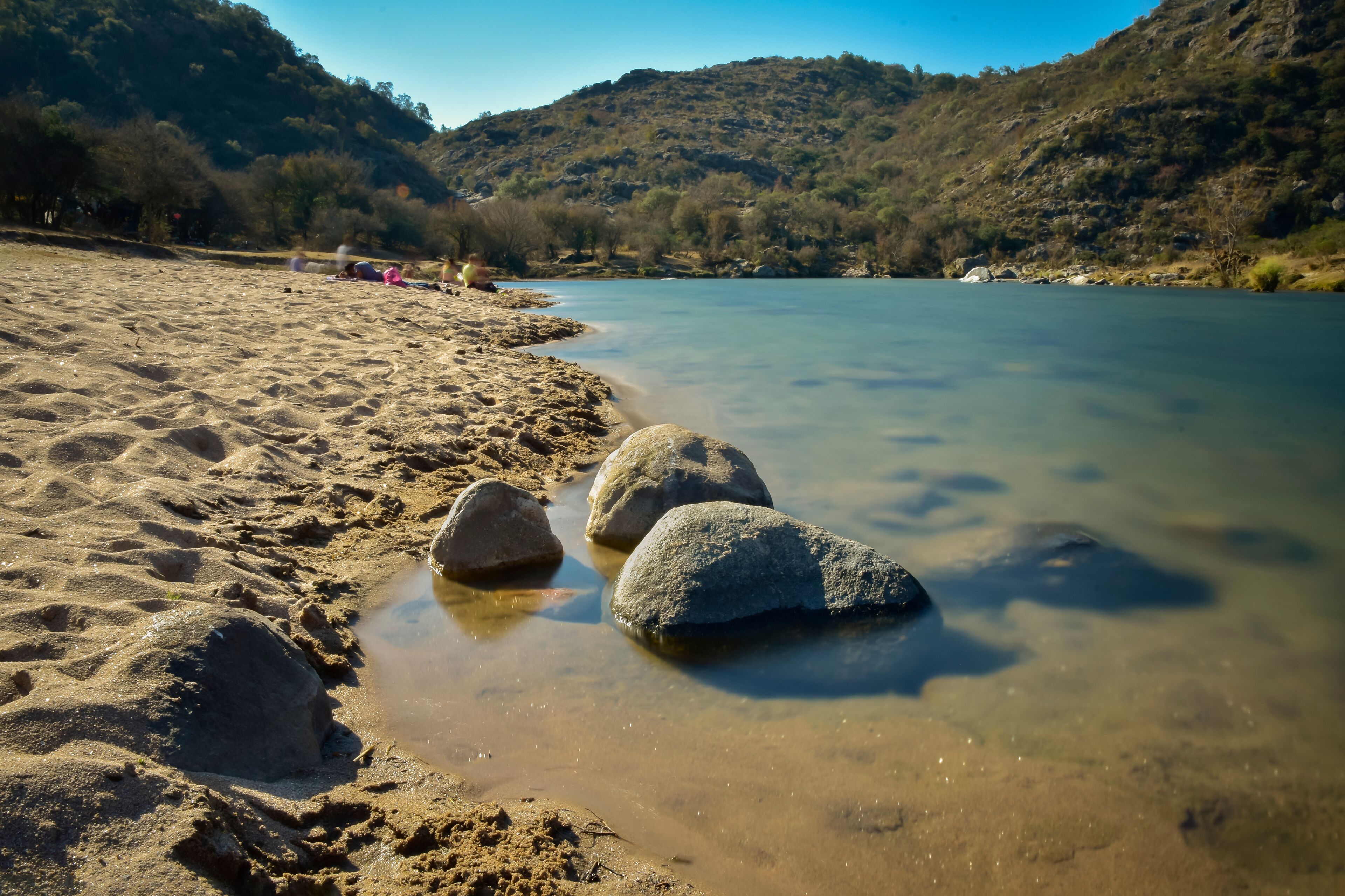 A riverbank with clear water, sandy shore, and large rocks.