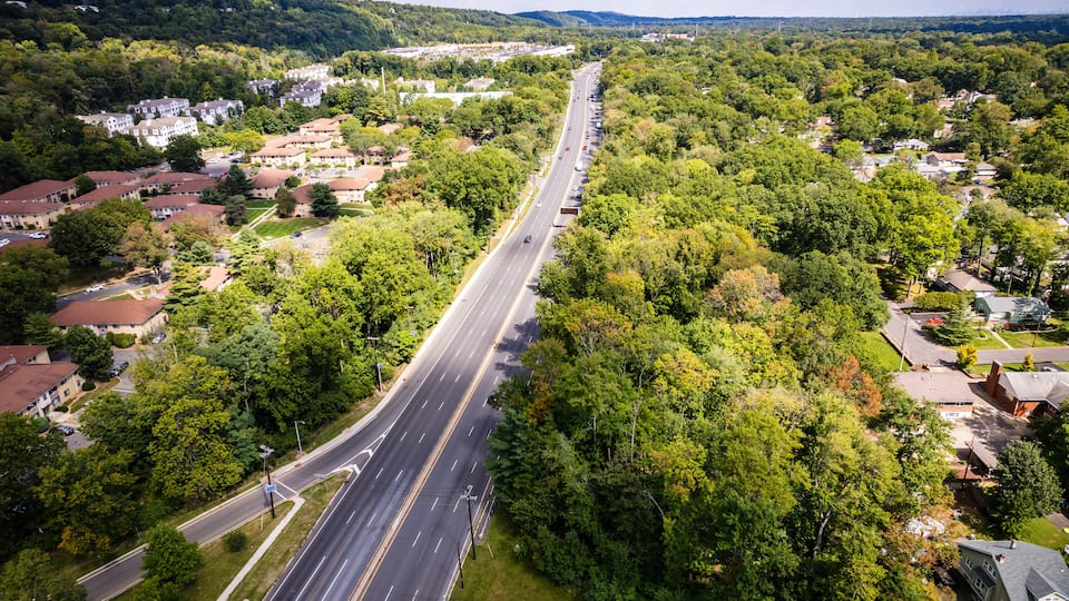 A highway cutting through a densely wooded area with residential buildings on either side.