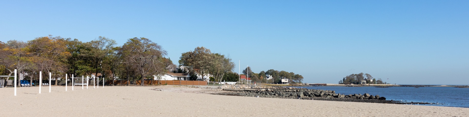 Une plage de sable fin, un ciel bleu dégagé, une digue rocheuse et des arbres en arrière-plan.