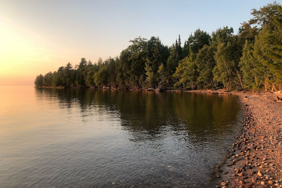 Un lago tranquilo al atardecer, con una orilla rocosa y árboles densos a lo largo de la costa.