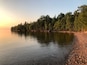 Ein ruhiger See bei Sonnenuntergang mit steiniger Uferlinie und dichter Baumgrenze.