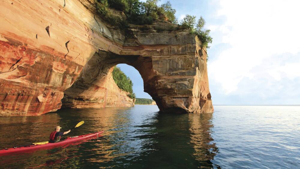 A kayaker in a red kayak under a natural rock arch over a body of water.