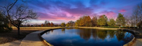 A serene park with a reflective pond, autumn-colored trees, and a clear sky.