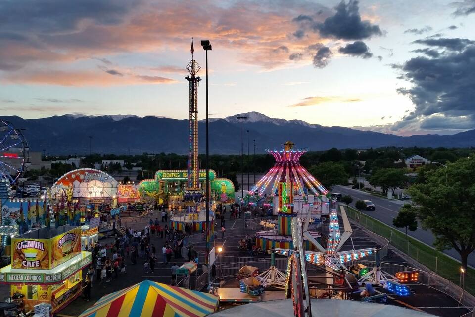 Um carnaval com atrações e barracas de comida, com montanhas e um céu ao pôr do sol ao fundo.