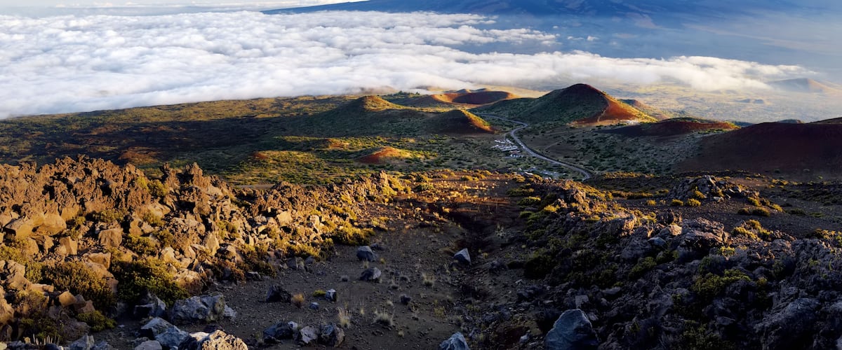 A mountainous landscape with a road winding through the hills, a cloud-covered valley below, and a clear sky above.