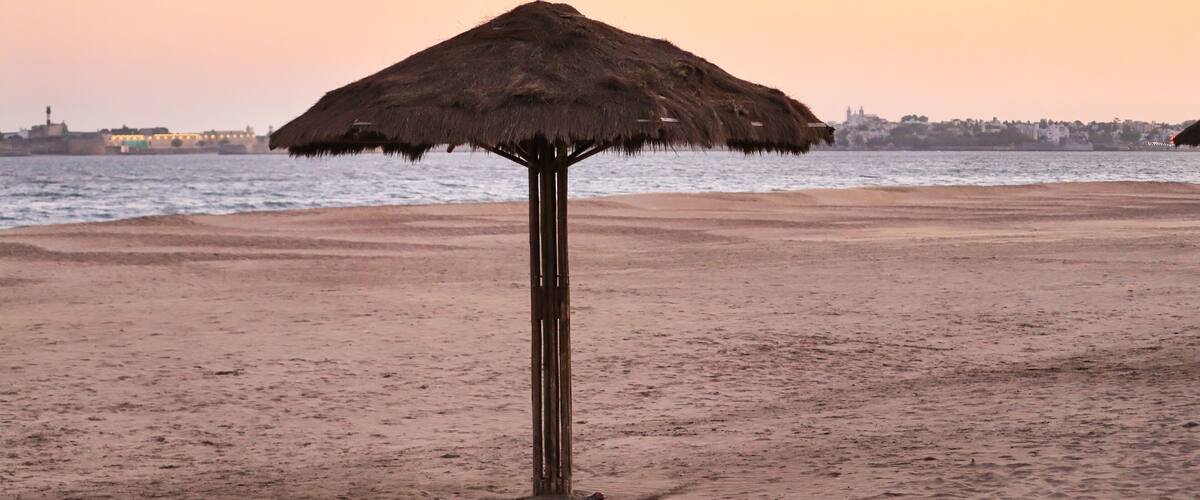 A beach with a thatched umbrella, sandy shore, and a distant cityscape.