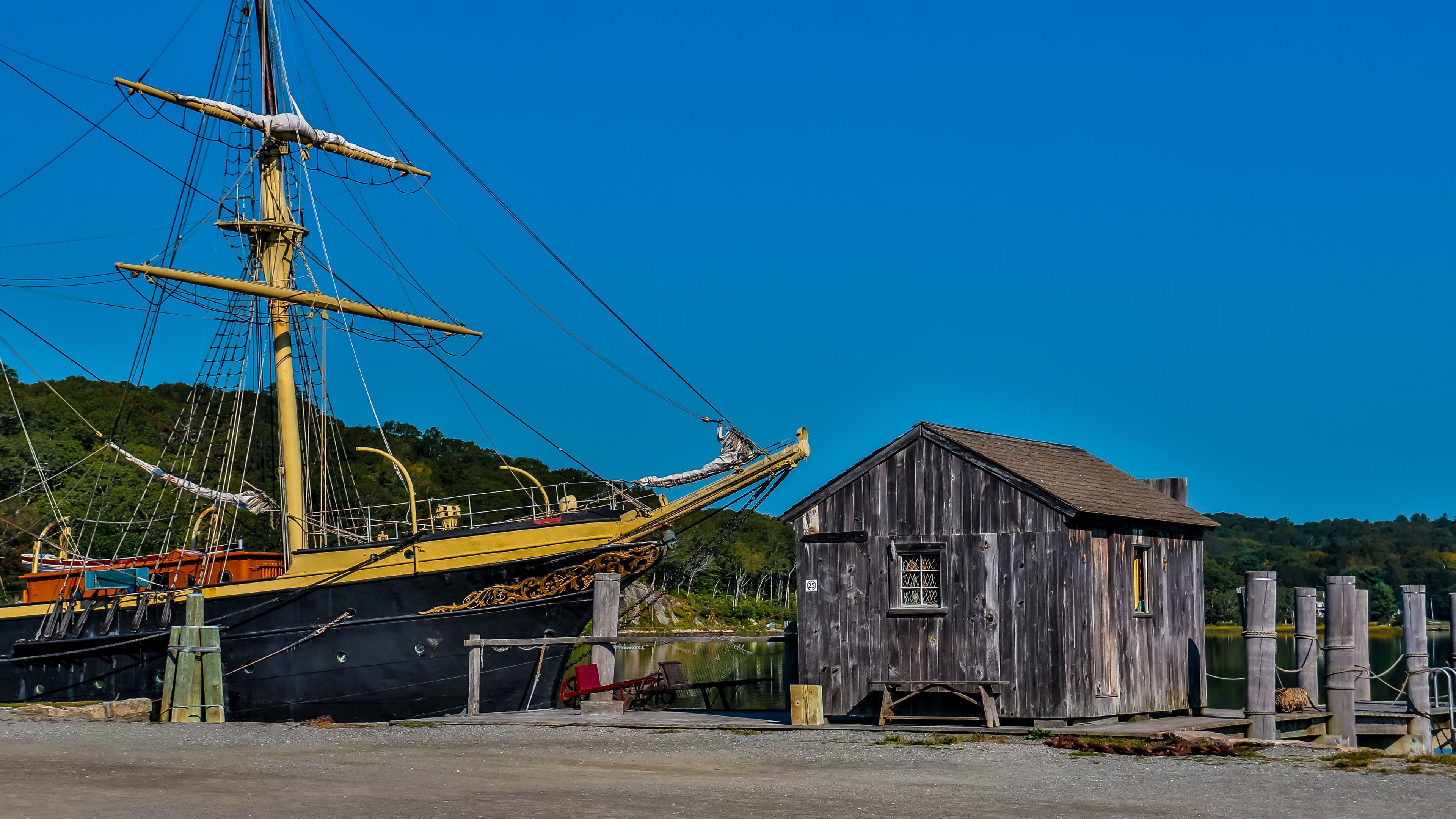 A tall ship docked at a pier, a wooden building, and a forested hill in the background.