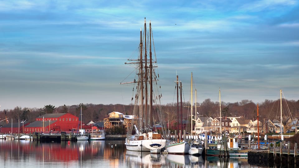 A harbor with several sailboats docked, a red building, and houses in the background.