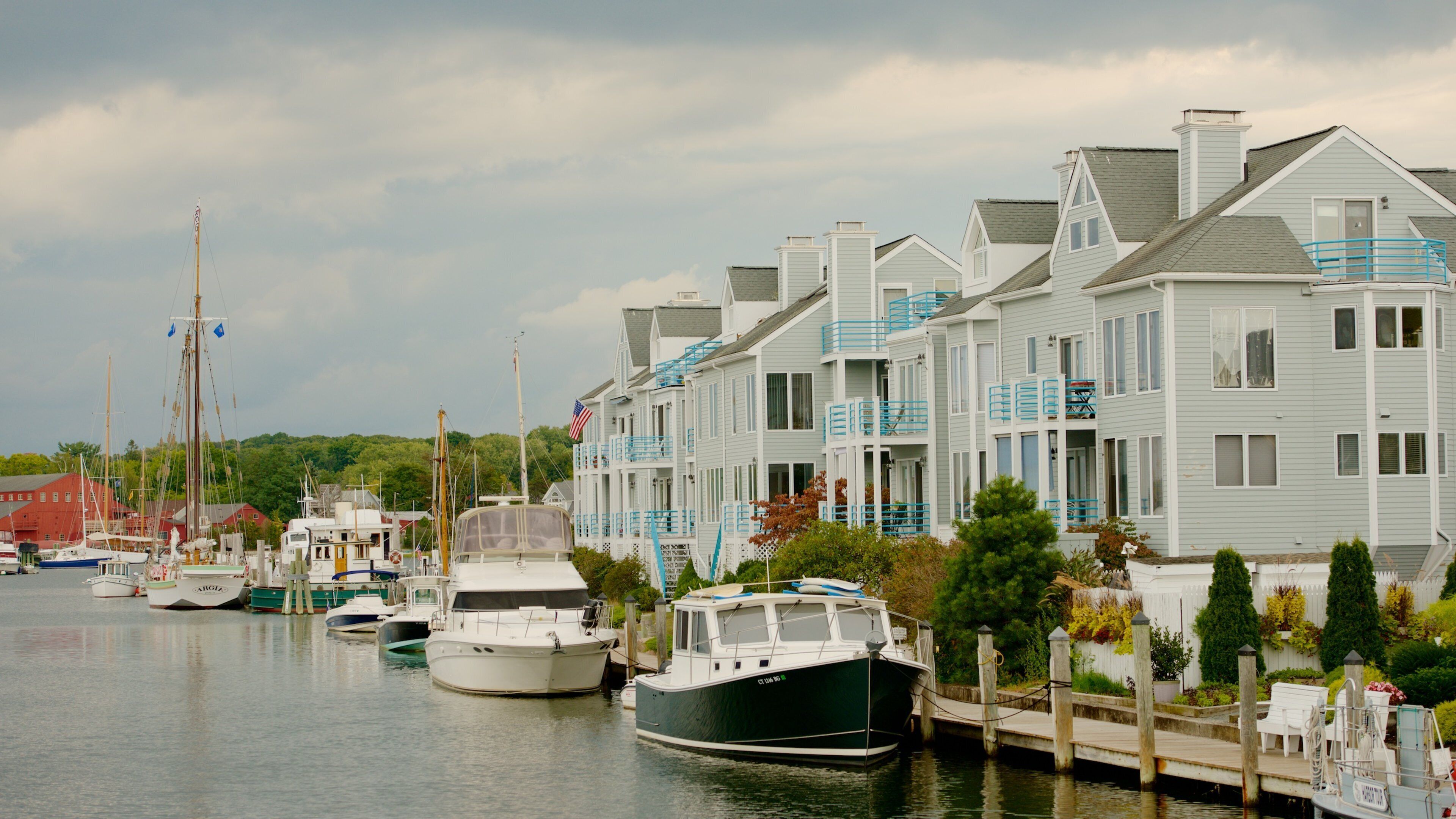 A marina with boats docked, a row of houses with blue railings, and an American flag on one of the boats.