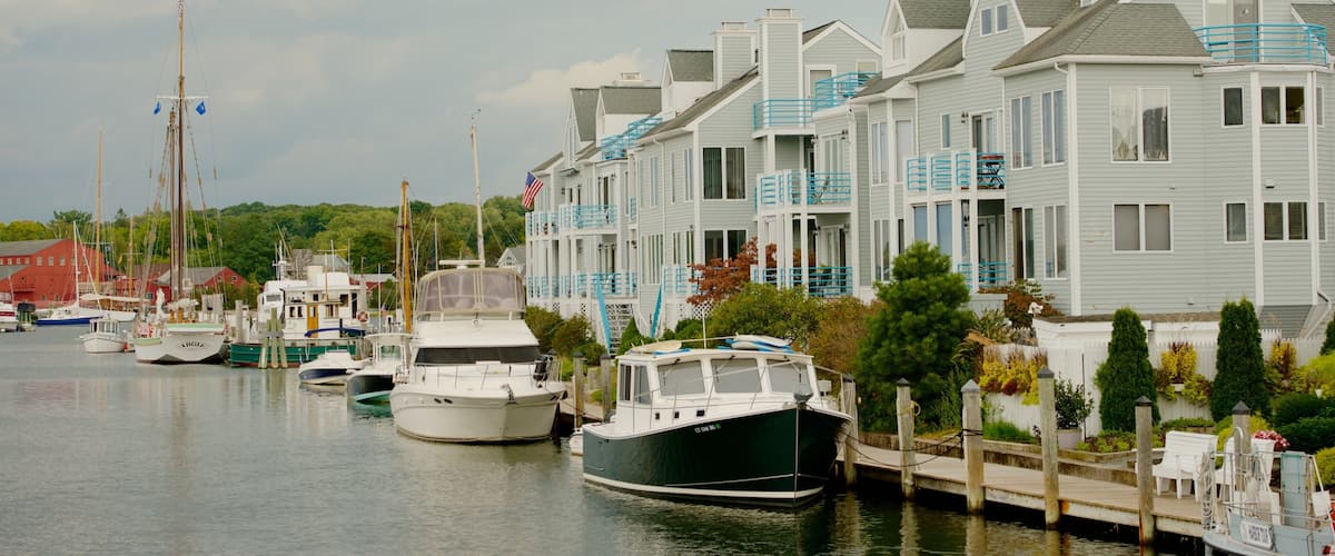 A marina with boats docked, a row of houses with blue railings, and an American flag on one of the boats.