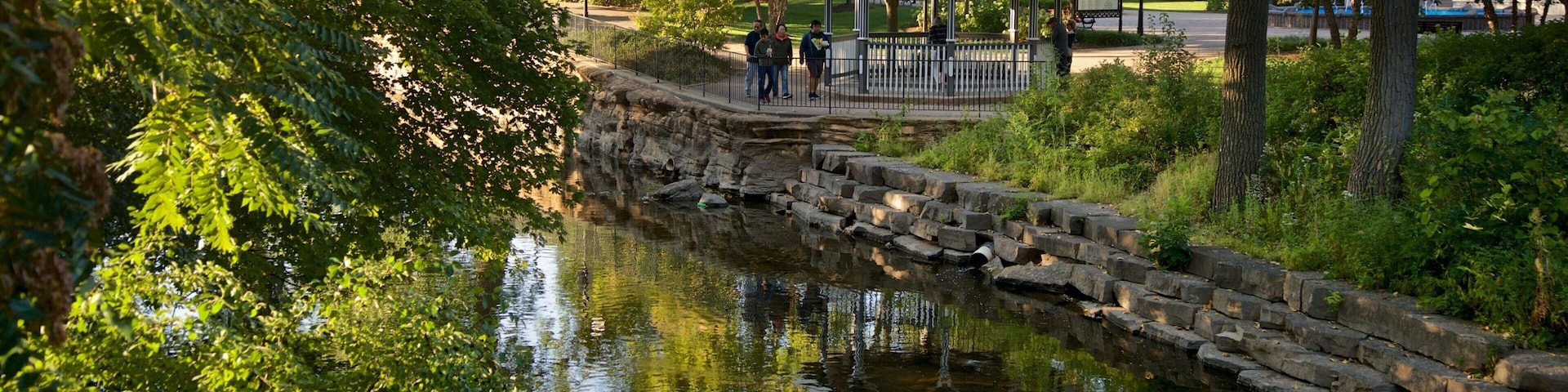A stone bridge over a calm river, surrounded by lush greenery and a gazebo in the background.