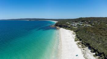 A coastal landscape with a sandy beach, clear turquoise waters, and a residential area with houses on a hillside.