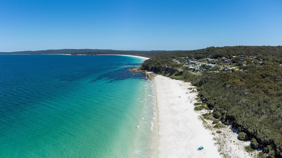 A coastal landscape with a sandy beach, clear turquoise waters, and a residential area with houses on a hillside.