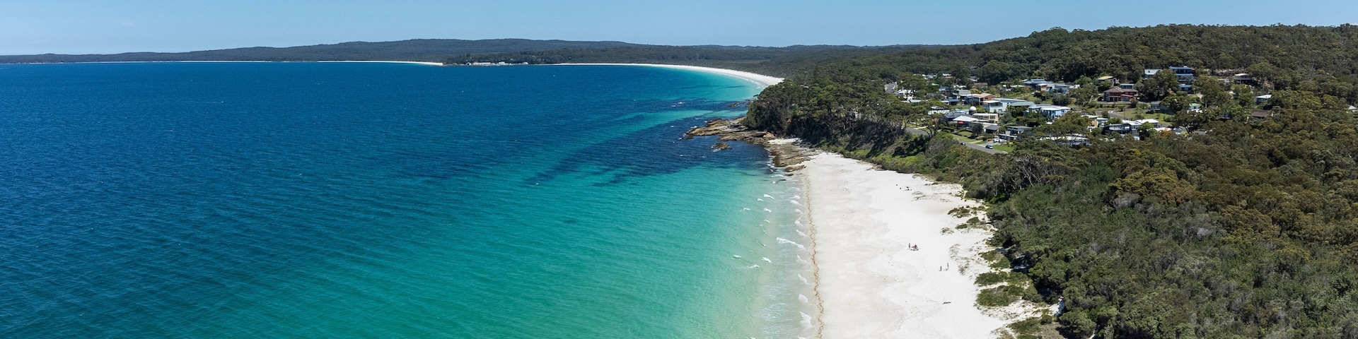 Paisagem costeira com praia arenosa, águas turquesa cristalinas e área residencial com casas numa encosta.
